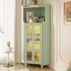 A green corner cabinet with glass doors is shown in a room setting, next to a window with sheer curtains and a small side table with a bust on it. Decorative items are on the shelves. Produktbild-Vorschau
