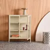 A beige cabinet with glass doors stands against a wood-paneled wall, with decorative objects on top and inside. A silver stool is to the right. Produktbild-Vorschau