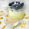 A jar of dessert topped with blueberries and powdered sugar, next to a small bowl of psyllium husk, surrounded by dried yellow flowers. Produktbild-Vorschau