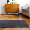 A dark gray rug is placed on a wooden floor in front of a wooden cabinet and a light-colored armchair. Anteprima dell’immagine prodotto