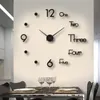 A large wall clock with numbers written out in words is mounted on a wall above a dining table set with plates and glasses. Produktbild-Vorschau