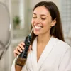 A woman in a white bathrobe is using a black and brown water flosser in a bathroom, smiling. Náhľad obrázku produktu
