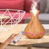 A hand holding a remote control is about to operate a wooden aroma diffuser that is emitting steam, placed on a wooden table next to a white wire basket. Produktbild-Vorschau
