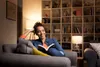 A woman is reading a book on a couch in a living room with bookshelves in the background. Produktbild-Vorschau