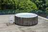 An inflatable hot tub and its control unit are placed on a wooden deck next to a swimming pool, with lush green trees in the background. Produktbild-Vorschau