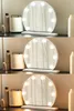 Three images of a round Hollywood-style makeup mirror with lights, displayed on a shelf with other cosmetic items. The lighting of the mirror changes between the three images. Podgląd zdjęcia produktu