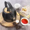 A black and gold kettle is placed on a wooden board, with a cup of tea, cookies, and books nearby. Náhled obrázku produktu