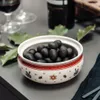 A bowl of black olives is placed on a gray surface, with a glass of red wine and a bottle in the background. Produktbild-Vorschau