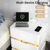A white nightstand with a tablet and phone charging on top, next to a couch. Náhľad obrázku produktu