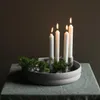 Four white candles are lit in a round, light gray candle holder with green foliage around the base, placed on a green tablecloth. Produktbild-Vorschau