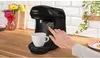 A hand presses a button on a black Bosch Tassimo coffee machine, with a cup of coffee below and flowers in the background. Produktbild-Vorschau