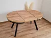 A wooden dining table with black legs is shown in a room with wooden floors and a white wall. Produktbild-Vorschau