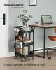A black rolling cart filled with office supplies and books is placed next to a desk with a laptop and a chair. Produktbild-Vorschau
