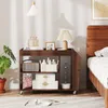 A wooden rolling cart with three shelves is placed next to a bed in a bedroom. The cart is decorated with various items, including a radio, baskets, boxes, and a lamp. Anteprima dell’immagine prodotto
