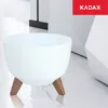 A white planter pot with wooden legs sits on a marble floor, with a geometric white structure and a red KADAX logo in the background. Náhled obrázku produktu