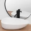 A white oval basin sink with black faucets is placed on a wooden surface, with a large round mirror reflecting the scene. Anteprima dell’immagine prodotto