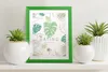 A green picture frame with a botanical print is displayed on a shelf between two potted plants. Produktbild-Vorschau