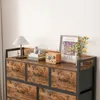 A close-up view of a rustic brown industrial dresser with seven drawers, featuring a wooden top and black metal frame. Decorative items like a lamp, books, a vintage radio, and a framed picture are placed on top. Náhled obrázku produktu