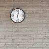 A round wall clock with a black frame and white face is mounted on a light gray brick wall. Voir les images du produit