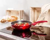A red frying pan with tomatoes sits on a black induction stove, with bread and wooden crates in the background. Podgląd zdjęcia produktu