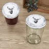 Two jars with lids decorated with a stag's head are placed on a wooden surface. One jar contains a dark red substance, possibly jam, while the other is empty. A wooden crate with blueberries and greenery is visible in the background. Podgląd zdjęcia produktu