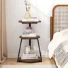 A three-tiered round side table with a wooden top and black metal legs, placed next to a bed in a bedroom setting. Podgląd zdjęcia produktu