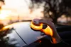 A hand holding a small, illuminated safety light above a car's roof during sunset. Produktbild-Vorschau