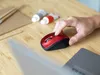 A hand is using a red wireless mouse on a wooden desk, with a laptop and art supplies in the background. Anteprima dell’immagine prodotto