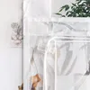 A close-up view of a white sheer curtain with a subtle leaf pattern, showing a clear plastic toggle and a basket of oranges in the background. Produktbild-Vorschau