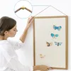 A woman is hanging a framed picture with butterflies on a white wall. A close-up inset shows how the frame is hung with a string and a brass hook. Produktbild-Vorschau
