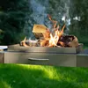 A close-up shot of a fire pit with logs burning, set on a metal stand with a drawer, outdoors on a grassy lawn with trees in the background. Podgląd zdjęcia produktu