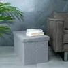 A gray fabric storage ottoman sits on a tiled floor next to a gray sofa, with a plant in the foreground and a gray tiled wall in the background. Produktbild-Vorschau