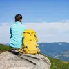 A woman with braided hair sits on a rock, looking out at a mountain landscape. A yellow backpack and trekking poles are beside her. Produktbild-Vorschau