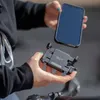 A person is holding a smartphone and a black clamp accessory, seemingly about to attach the phone to the clamp. Produktbild-Vorschau