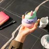 A hand holding a colorful unicorn poop squishy toy on a dark marble table with a notebook and pen. Voir les images du produit