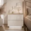A modern white nightstand with gold accents and LED lighting, placed next to a bed in a bedroom. Produktbild-Vorschau