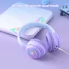 A pair of purple gaming headphones with cat ears and LED lights, placed on a white surface next to a gaming controller. Podgląd zdjęcia produktu