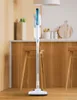 A white and blue stick vacuum cleaner is standing upright in a kitchen setting with wooden cabinets and stools. Náhled obrázku produktu
