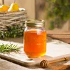 A jar of honey with a honey dipper on a wooden board, with a basket of pears and greenery in the background. Voir les images du produit
