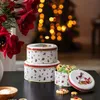 Christmas-themed cookie tins are stacked on a dark surface, with a bouquet of red flowers and a blurred Christmas tree in the background. Produktbild-Vorschau