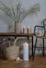 A tall, white LED candle with a lit flame sits on a wooden floor next to a woven basket filled with bottles and a fluffy brown item. Behind the basket is a wooden bench with a vase of dried grass and two glasses. Produktbild-Vorschau