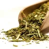 A wooden scoop filled with dried green tea leaves and some straw-like pieces, with some spilling onto a white surface. Produktbild-Vorschau