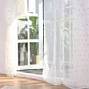 A sheer curtain with pom-poms hanging in front of a window with plants visible outside. Produktbild-Vorschau