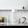 A white soda maker sits on a kitchen counter with two bottles of water and a glass of water with rosemary. Produktbild-Vorschau