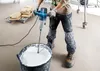 A person is mixing a white substance in a black bucket with a blue and silver power mixer. Produktbild-Vorschau