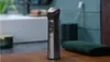 A Philips hair clipper is placed on a wooden surface next to a green towel. The background shows a bathroom setting with a sink and tiled wall. Anteprima dell’immagine prodotto