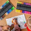 A child's hands are coloring in a book with markers. Two sets of markers are laid out on a wooden table. Produktbild-Vorschau