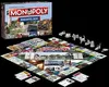 A Monopoly board game box and board are displayed on a black background, along with game pieces. Produktbild-Vorschau