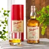 A bottle of Glenfarclas Highland Single Malt Scotch Whisky, a glass of whisky, and its red presentation tube are arranged on a wooden surface with greenery in the background. Produktbild-Vorschau