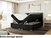 A bedroom scene featuring a black upholstered bed with its storage compartment open, revealing the mattress and topper. The room has soft lighting and a neutral color palette. Produktbild-Vorschau